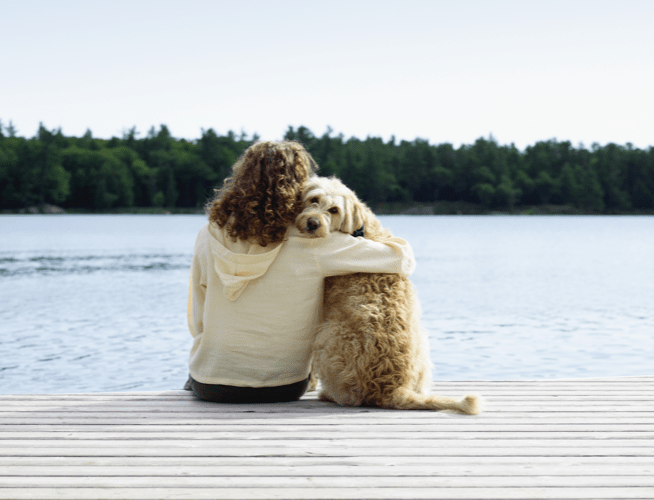 Candid snapshot of a woman with her arm around her pet dog as they sit together on a lakefront dock